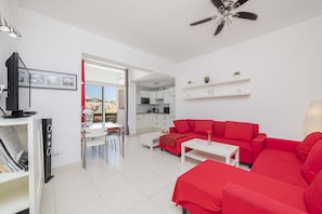 Uma sala de estar moderna com um sofá vermelho, uma mesa de centro branca e uma TV em um móvel. Há uma área de jantar com mesa e cadeiras, e uma cozinha ao fundo.