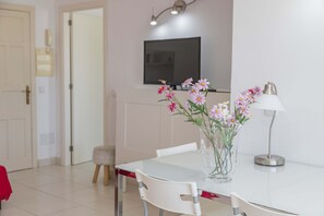 Uma área de refeições com uma mesa branca, cadeiras, um vaso com flores, um abajur e uma televisão fixada na parede.