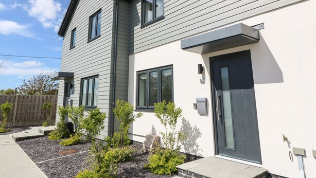 A modern two-story house with a grey exterior, a dark front door, and a small garden area with young trees and shrubs. - Dune Retreat (Ty Croes)