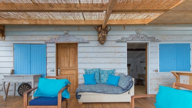 Tunnelmallinen, maalaishenkinen puumökki, jossa on olkikatto, puinen penkki ja sinisillä tyynyillä varustettu sohva.