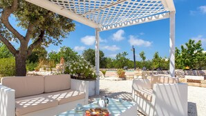 Ein Sitzbereich im Freien mit einem weißen Korbsessel und Tisch, einer Pergola darüber und Blick auf den umliegenden Garten und Poolbereich.