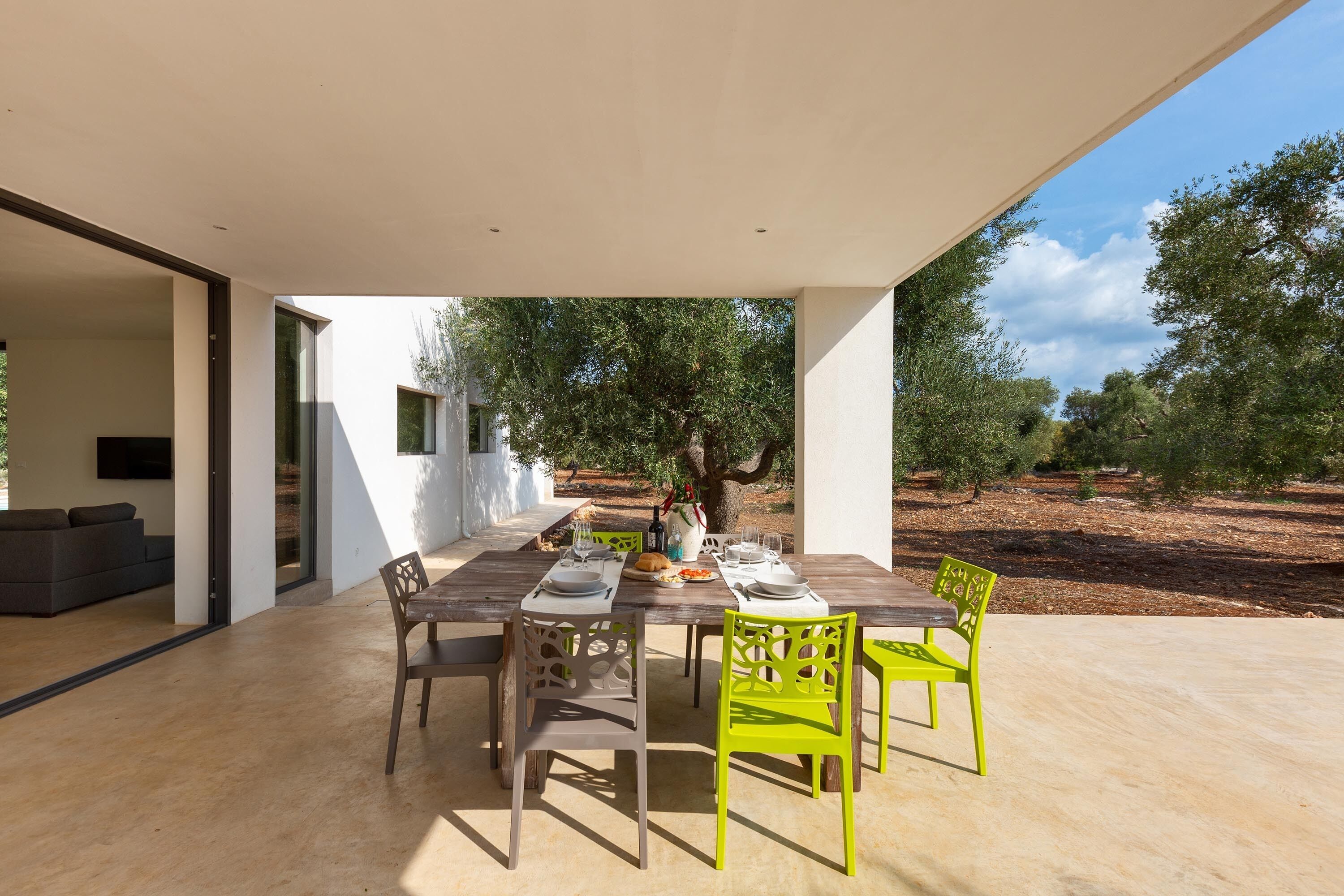 Un área de comedor al aire libre con una mesa de madera preparada para dos personas, rodeada de sillas y con vistas a olivos al fondo.