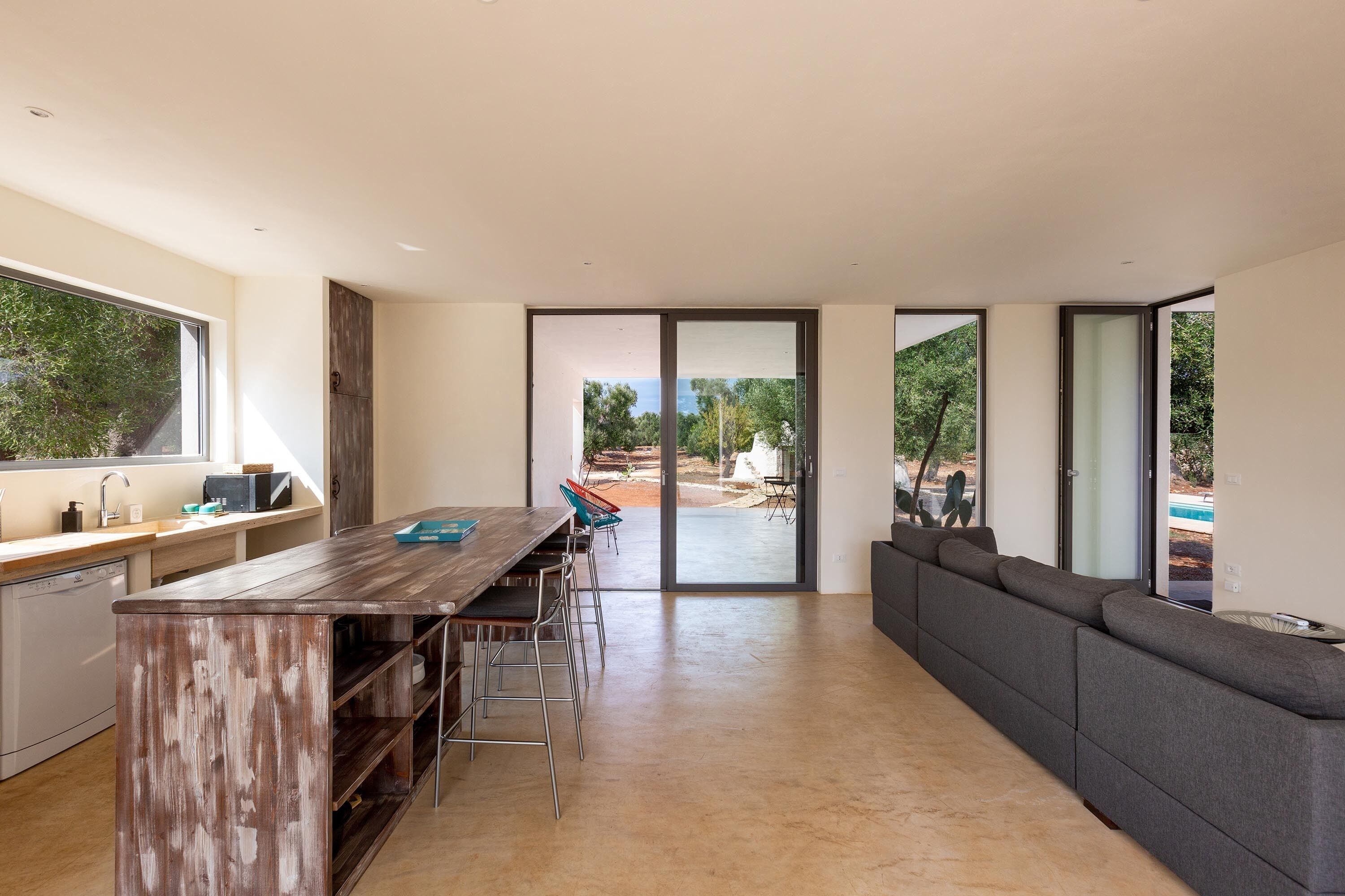 A modern kitchen with a wooden island, a dining area with chairs, and a large grey sofa.