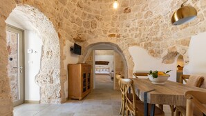 A rustic stone interior with a wooden dining table and chairs, a kitchen area, and a visible bed in the background. - 2220 Trulli Biancapetra by Perle di Puglia (Francavilla Fontana)