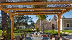 An outdoor dining area with a pergola, wicker chairs, a stone table, and a view of a stone building in the background. - 2220 Trulli Biancapetra by Perle di Puglia (Francavilla Fontana)