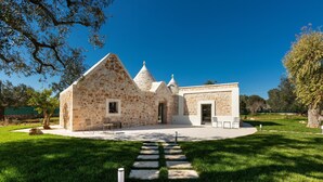 A stone building with a stone pathway leading to a door, surrounded by grass and trees. - 2220 Trulli Biancapetra by Perle di Puglia (Francavilla Fontana)