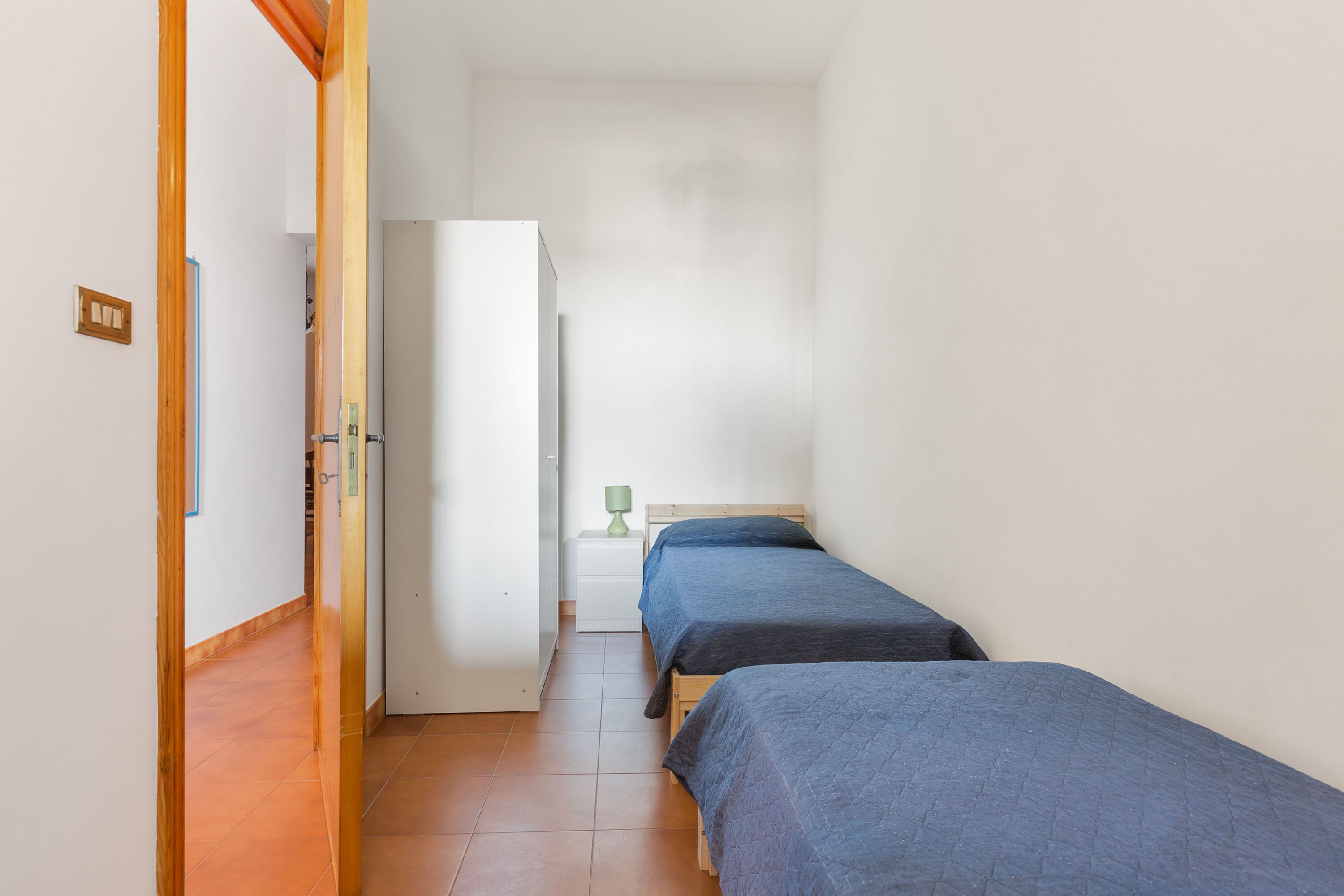 A narrow hallway with two beds, a white wardrobe, and a small bedside table with a lamp.