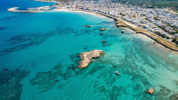 An aerial view of a coastal town with a clear beach, rocky outcrops, and a marina. - 2191 Appartamento Blue Dreams by Barbarhouse (Melendugno)