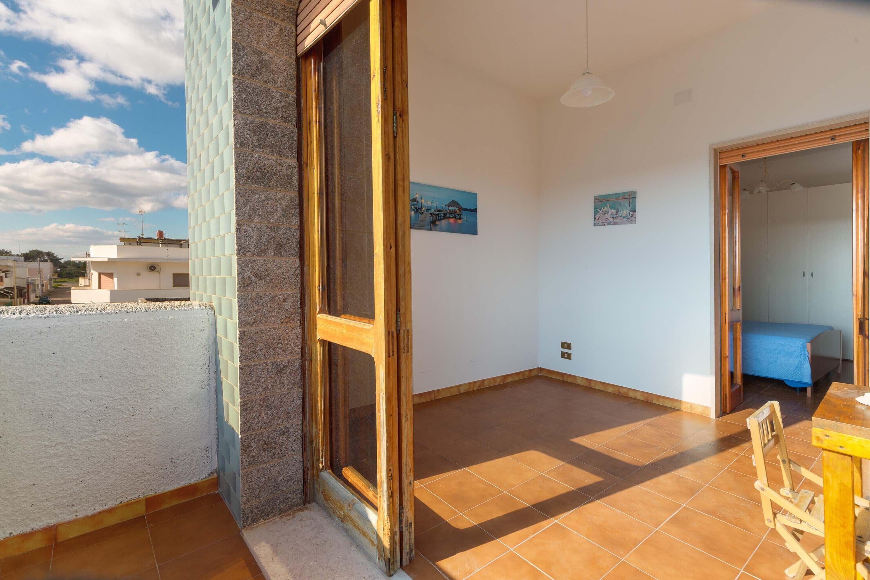 A room with a large window, a wooden door, a tiled floor, and a bed with a blue cover.