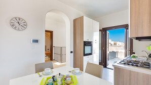 Une cuisine moderne dotée d’une cuisinière intégrée, un coin repas avec une table dressée pour deux personnes, et une vue sur la ville par la porte ouverte.
