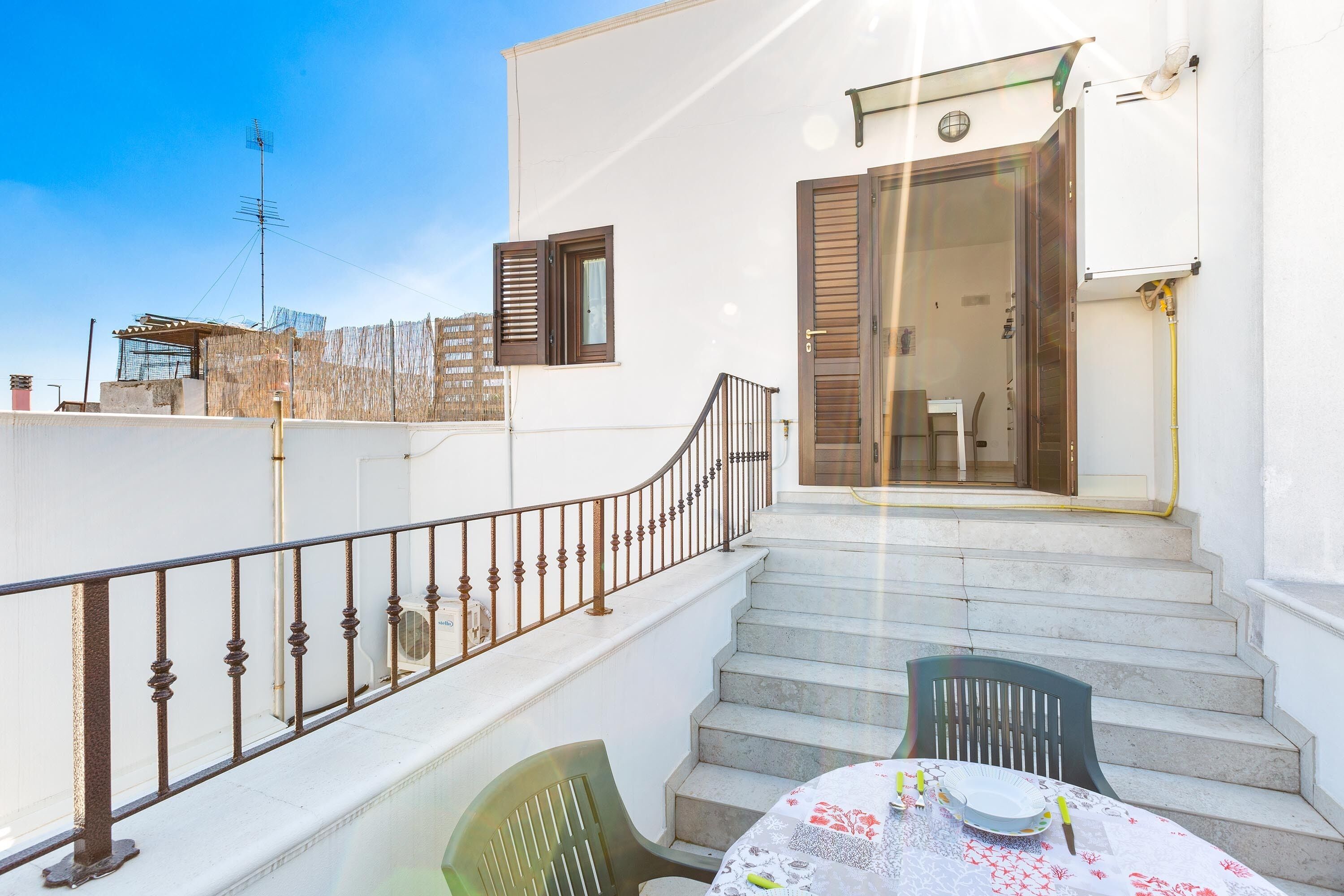 Un balcone con tavolo e sedie, una scala che conduce a una porta aperta e vista sulla zona circostante.