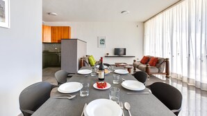 A modern dining area with a grey table set for four, a kitchen in the background, and a living area with a sofa and a TV. - 2178 Appartamento Negroamaro by Barbarhouse (Leverano)