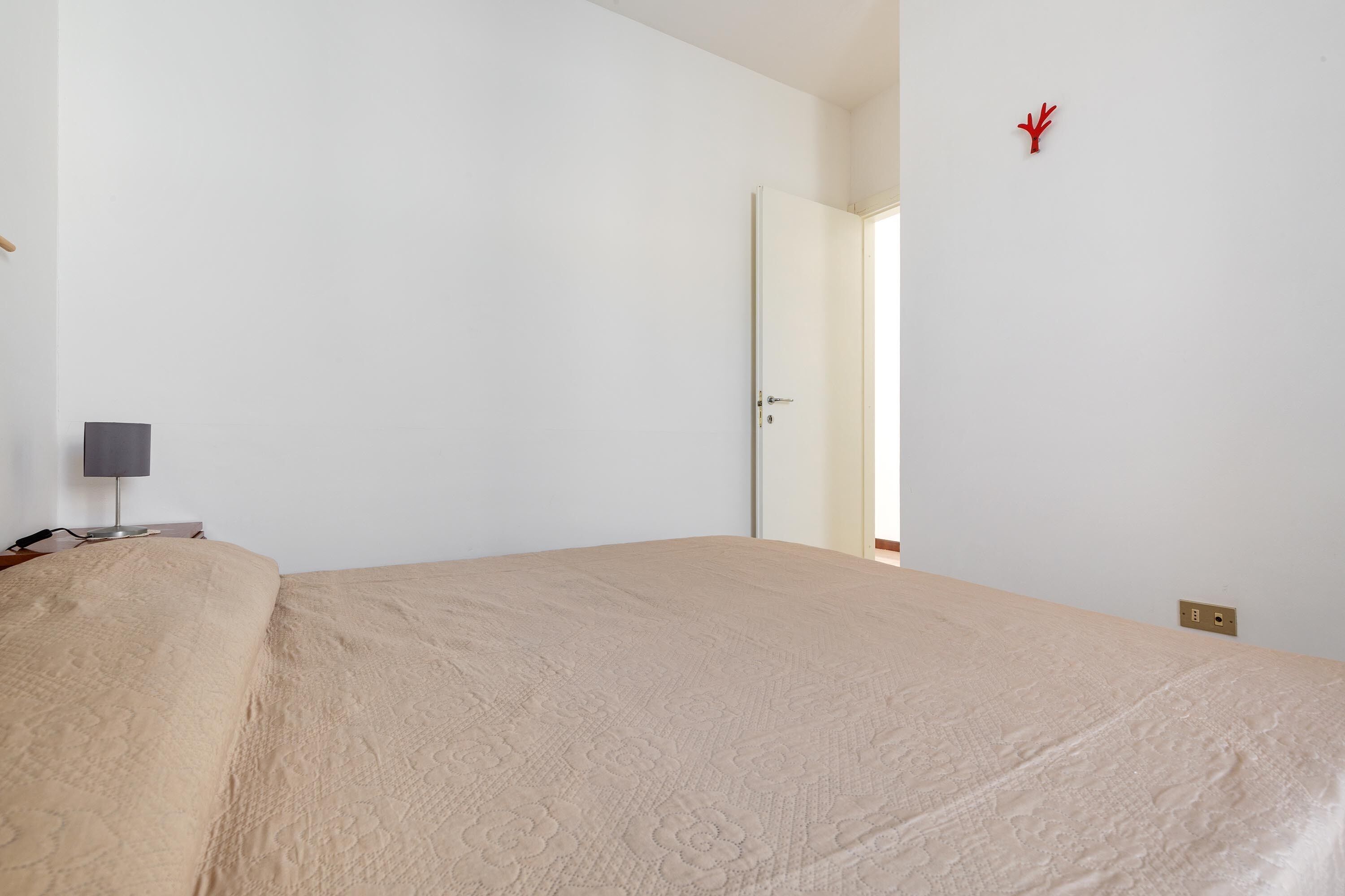 A room with a bed, a bedside table with a lamp, and a door leading to another room.