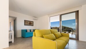 Ein modernes Wohnzimmer mit einem gelben Sofa, einem blauen Sessel und einer großen Schiebeglasdoor, die auf einen Balkon mit Meerblick führt.