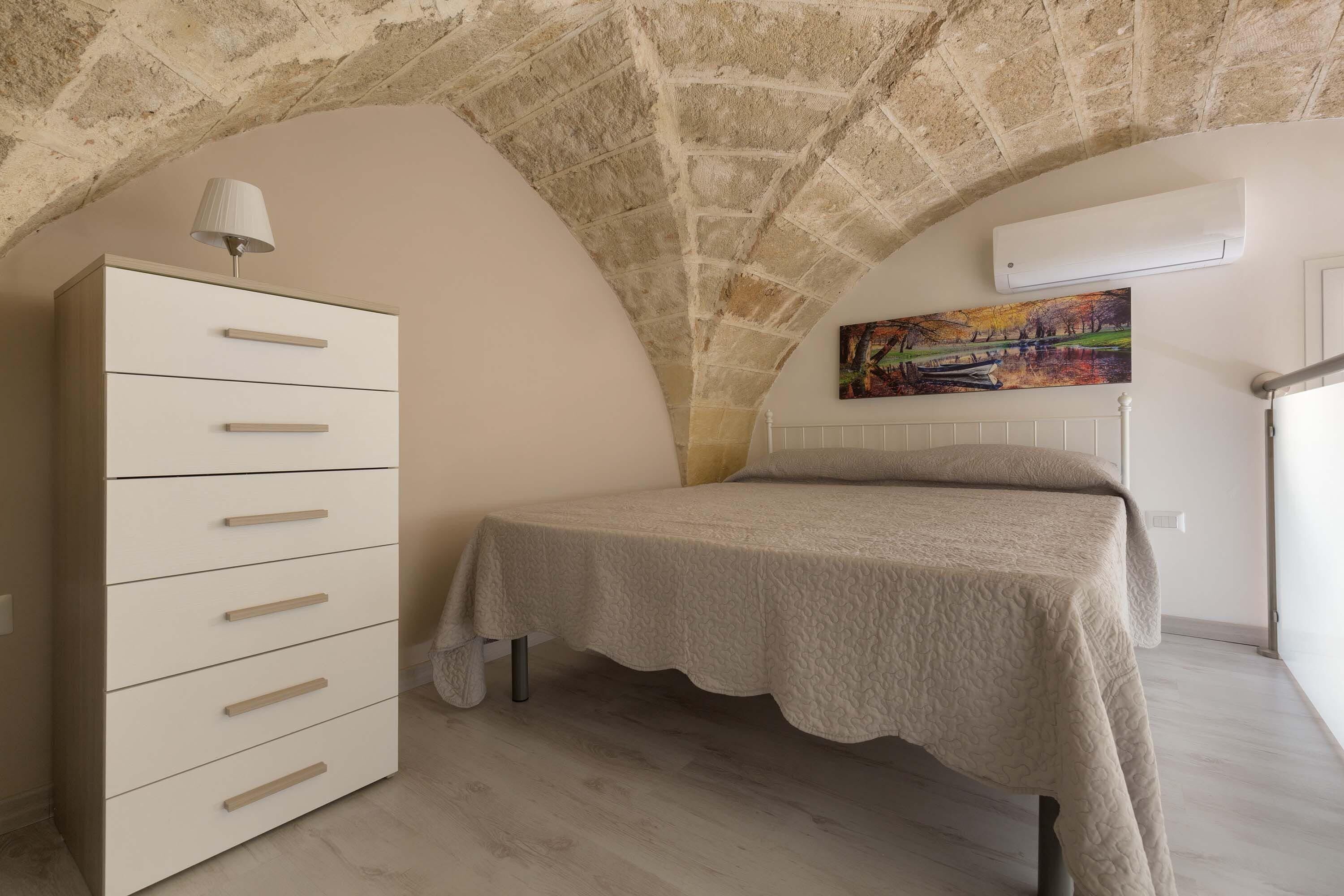 Een slaapkamer met een bed, een kaptafel, een schilderij aan de muur en een stenen plafond.
