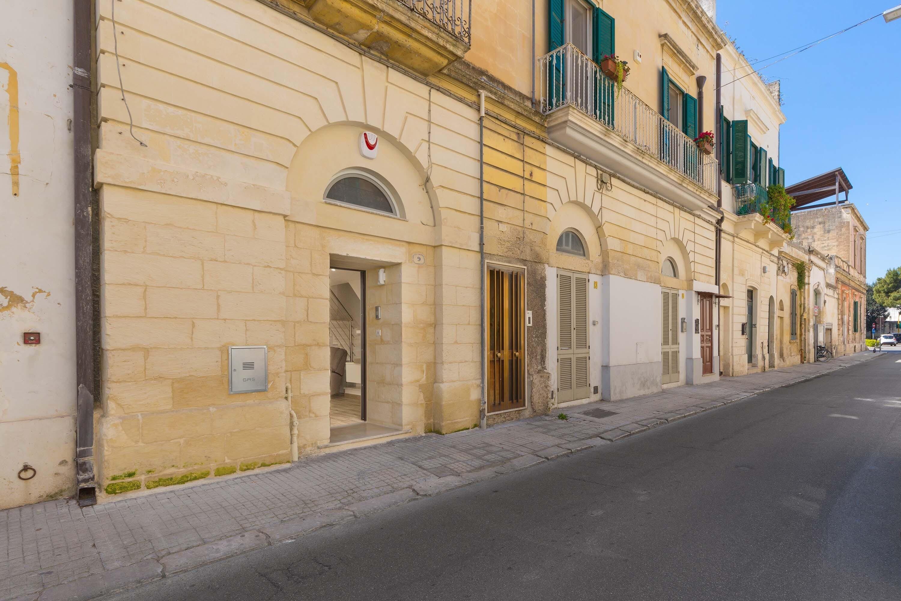 A row of historic buildings with arched doorways and balconies, a clear sky, and a quiet street.