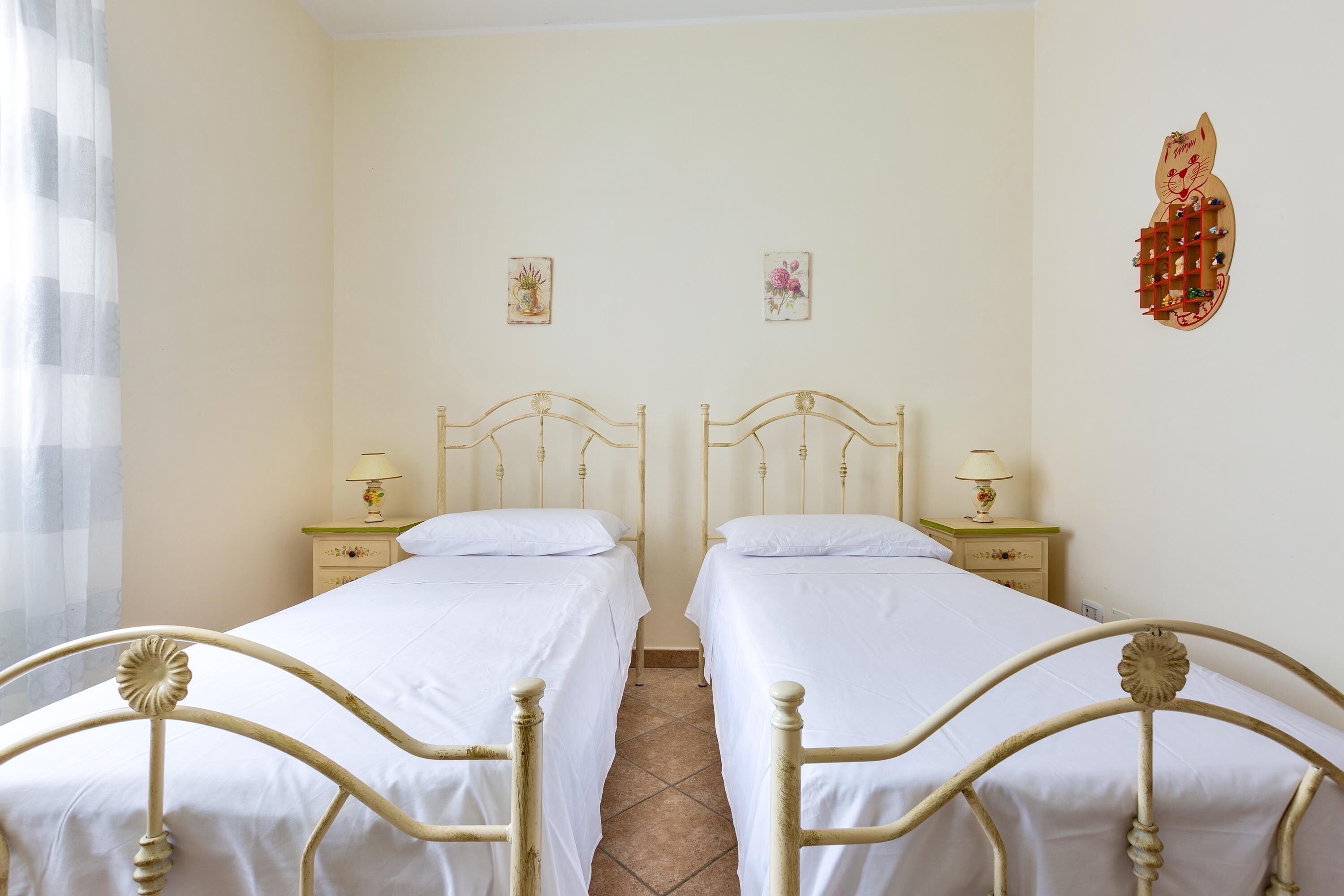 Une chambre avec deux lits simples, chacun doté d’une table de chevet et d’une lampe. Les murs sont de couleur unie, et une décoration murale orne l’espace au-dessus des lits.