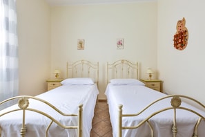 A room with two single beds, each with a nightstand and a lamp. The walls are plain, and there is a decorative wall hanging above the beds.