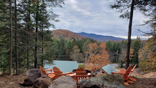A rustic cabin with a blue roof surrounded by a forest, with several wooden chairs and large rocks in the foreground.