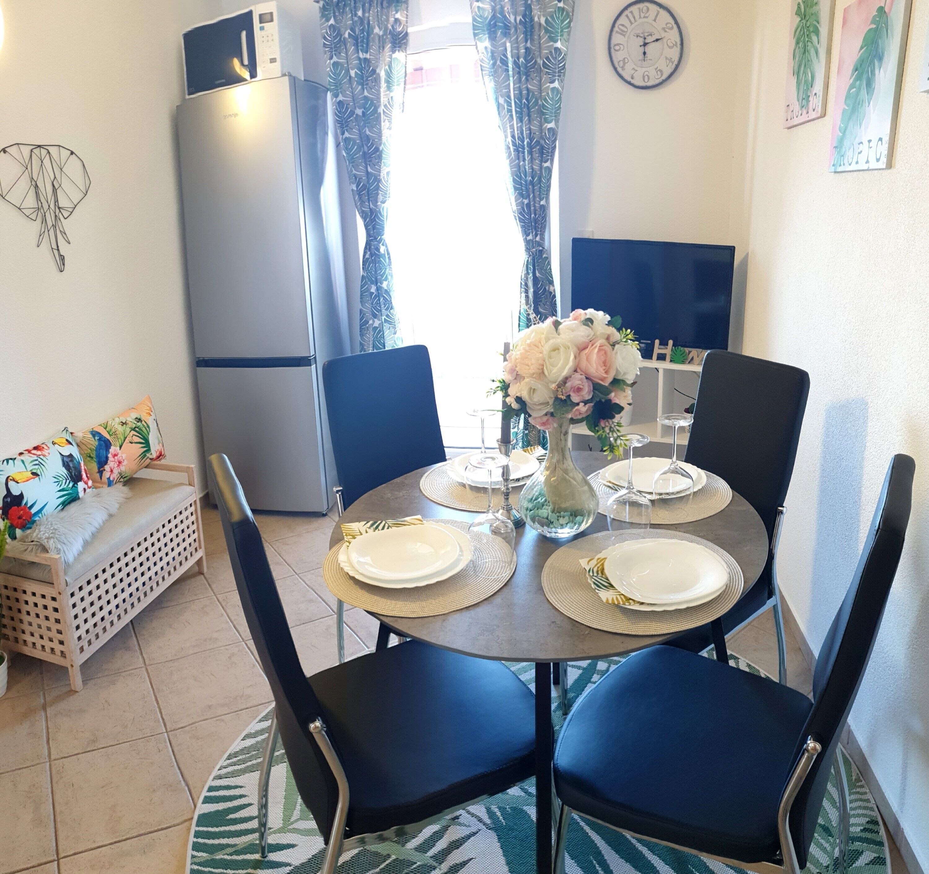 A dining area with a round table set for four, surrounded by chairs. A refrigerator is visible in the background, and a clock is mounted on the wall.