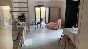 Una habitación con cama, una mesa con silla, una zona de cocina con lavabo y un ventanal corredero que da a un espacio exterior.