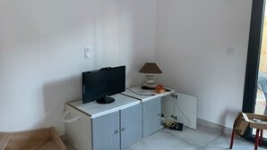 Una habitación con una televisión de pantalla plana sobre un mueble blanco, una lámpara y una silla cerca de un ventanal corredero.