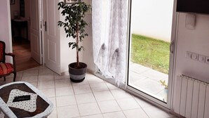 Ein geräumiger Raum mit einem großen Schiebeglastür, die zu einer Rasenfläche führt, einer Zimmerpflanze, einem Tisch mit einer Fernbedienung und einer Deckenleuchte.