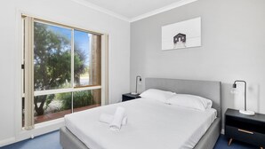 A modern bedroom with a large window, a bed with white linens, two bedside tables with lamps, and a framed picture on the wall. - Esplanade Townhouse 4-209 Only 50m to Beach (Torquay)