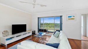 A modern living room with a flat-screen TV, white sofa, and a large window offering a view of greenery. - Esplanade Townhouse 4-209 Only 50m to Beach (Torquay)