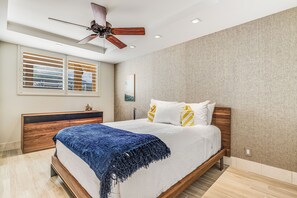 A bedroom with a bed, wooden headboard, ceiling fan, window blinds, and a view of the outside.