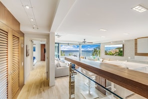 Una cocina moderna con una isla grande, vista al océano y un área de comedor con barandilla de vidrio.