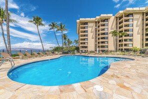 A swimming pool with lounge chairs and palm trees, surrounded by a multi-story building with balconies.