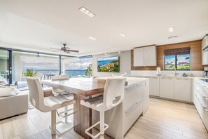 A modern kitchen with a large island, white bar stools, and a view of the ocean.