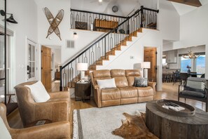Ein geräumiges Wohnzimmer mit einer Ledersofa, einem Couchtisch und einem rustikalen Holznachttisch. Eine Treppe mit schwarzer Geländer führt zu einer oberen Ebene.
