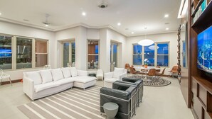 A modern living room with a white sectional sofa, a black leather ottoman, a dining area with a round table and chairs, and a large flat-screen TV mounted on the wall. - Ocean View, Modern Luxury w/Jacuzzi & access to all amenities! (Puntarenas)