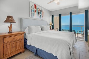A bedroom with a bed, a nightstand, a lamp, a painting, and a view of the ocean.