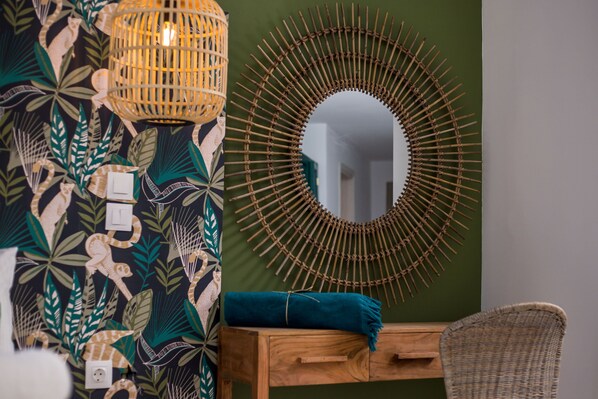 A room with a green wall, a woven light fixture, a circular mirror, a wooden dresser, and a wicker chair. - Marini Seaside (Rhodes)