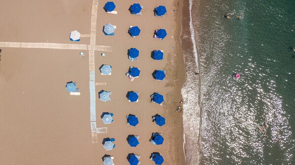 An aerial view of a beach with rows of blue umbrellas and beach chairs, a sandy shore, and the ocean. - Marini Seaside (Rhodes)