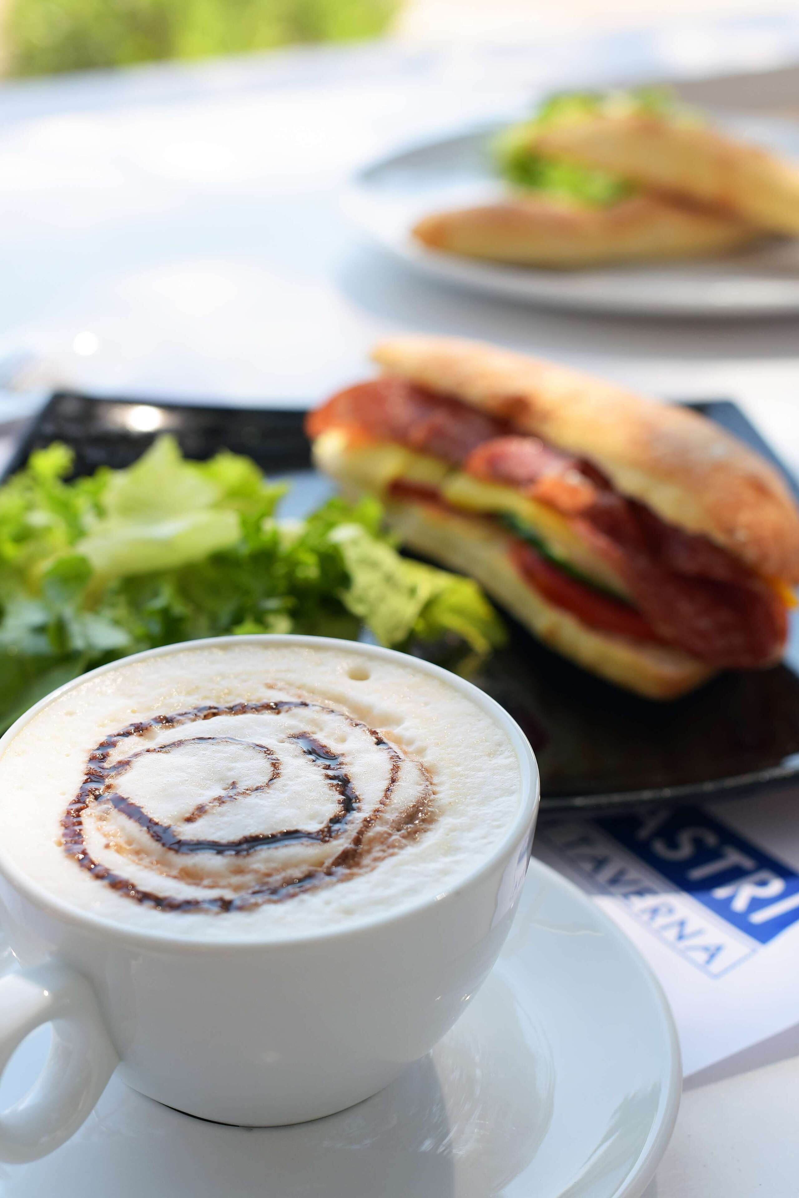 Een kop koffie met latte-art op een schotel, een belegd broodje en een bord met groene groenten.