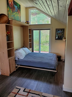 A bedroom with a bed, wooden shelving, a large window, and a door leading outside.