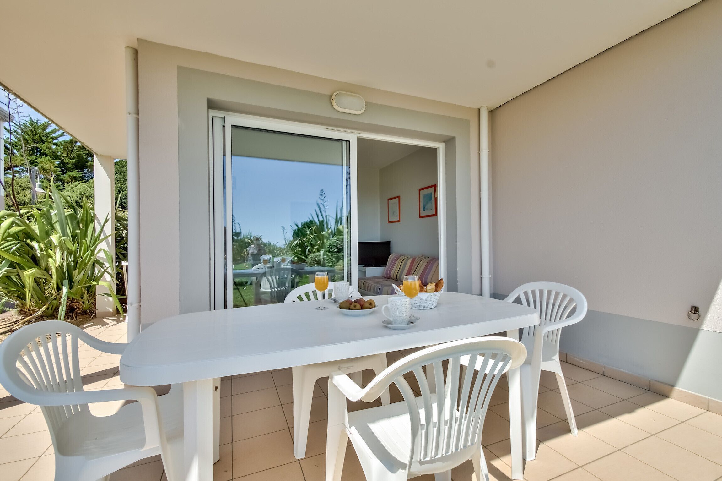 A white table and chairs on a balcony with a glass door, a breakfast setup with orange juice, and a view of greenery and a building in the background.