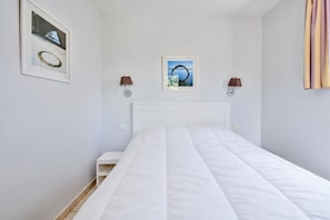 A bedroom with a white bed, a nightstand, two wall-mounted lamps, and two framed pictures on the wall.