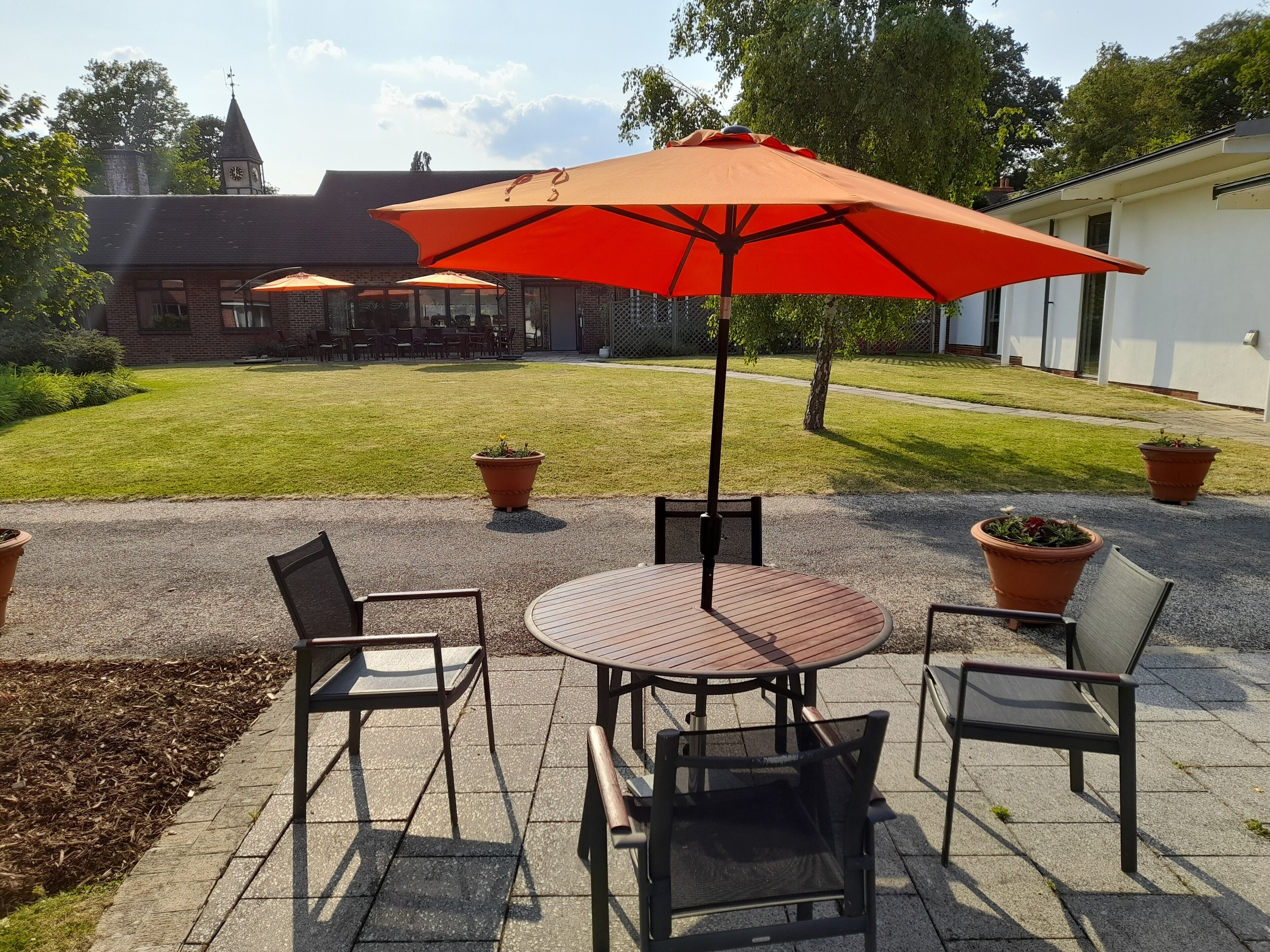 Un espace patio comprenant une table ronde et quatre chaises, un grand parasol rouge et des plantes en pot.