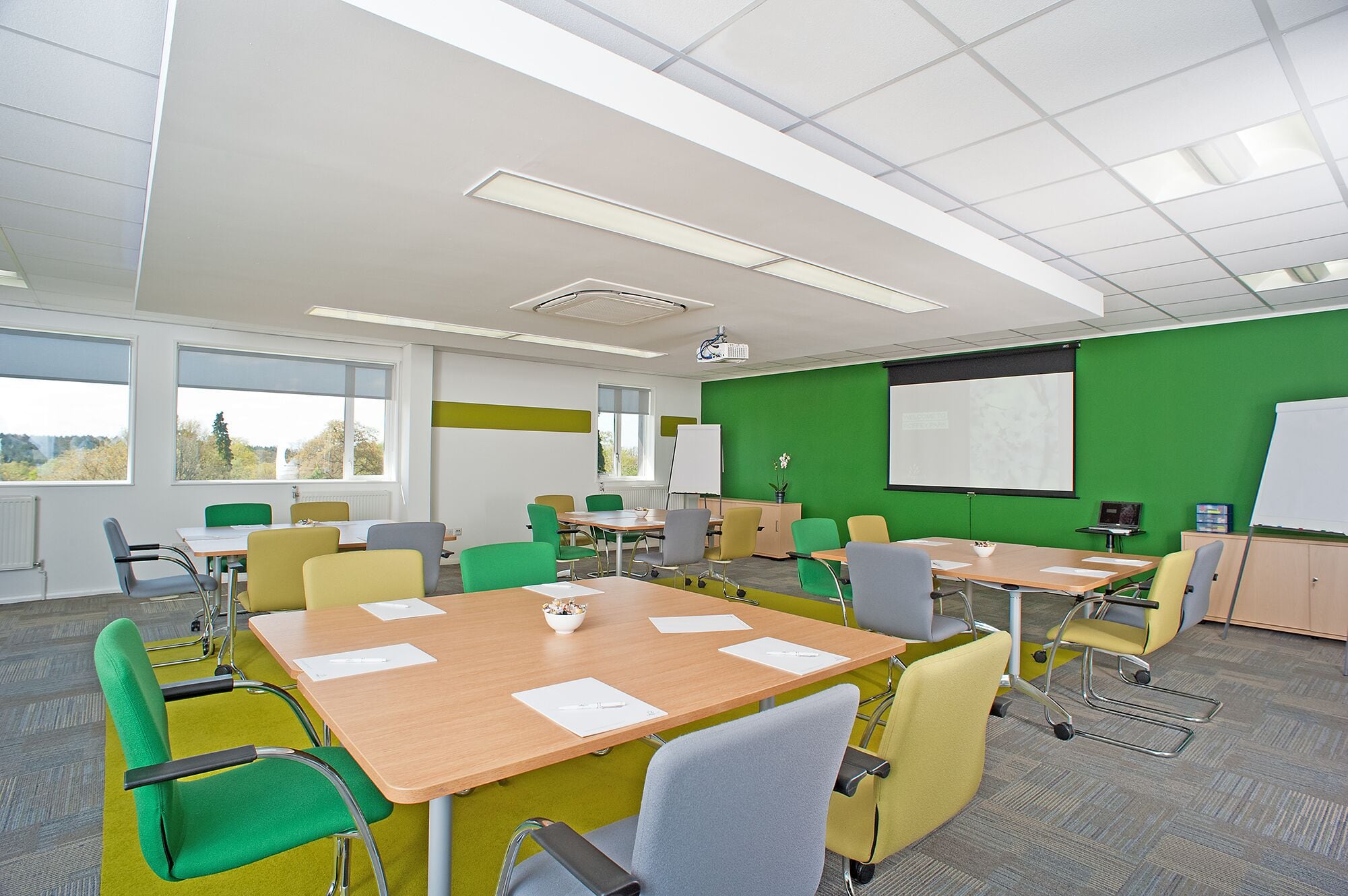 A conference room with multiple tables and chairs, a projector, a whiteboard, and a green screen wall.
