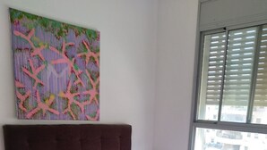 A bedroom with a bed, a window with blinds, a wall-mounted painting, and a small bedside table.