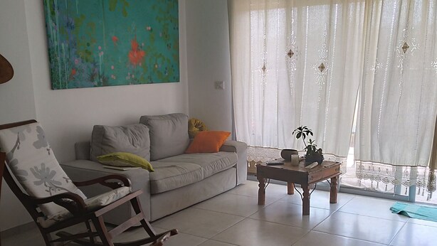 A living room with a sofa, a rocking chair, a coffee table, and a large window with sheer curtains.