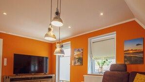 A living room with orange walls, a flat-screen TV, a wooden entertainment unit, and a window with a view of the outdoors.