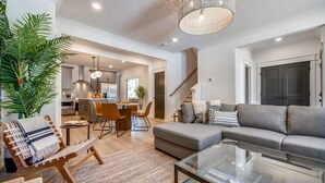 A modern living room with a grey sofa, a glass coffee table, and a woven chair. There is a staircase leading to another level, a kitchen area in the background, and a large plant on the left. - Beautiful Luxury Renovation, 5 minutes to Downtown (Nashville)