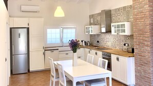 A modern kitchen with white cabinetry, a brick accent wall, and a dining area with a white table and chairs. - Bio Resort Mediterraneo (Sant Martí Sarroca)