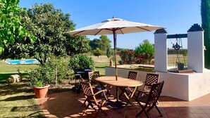 An outdoor patio with a round table and chairs, a large umbrella, and a view of a swimming pool and garden. - Bio Resort Mediterraneo (Sant Martí Sarroca)